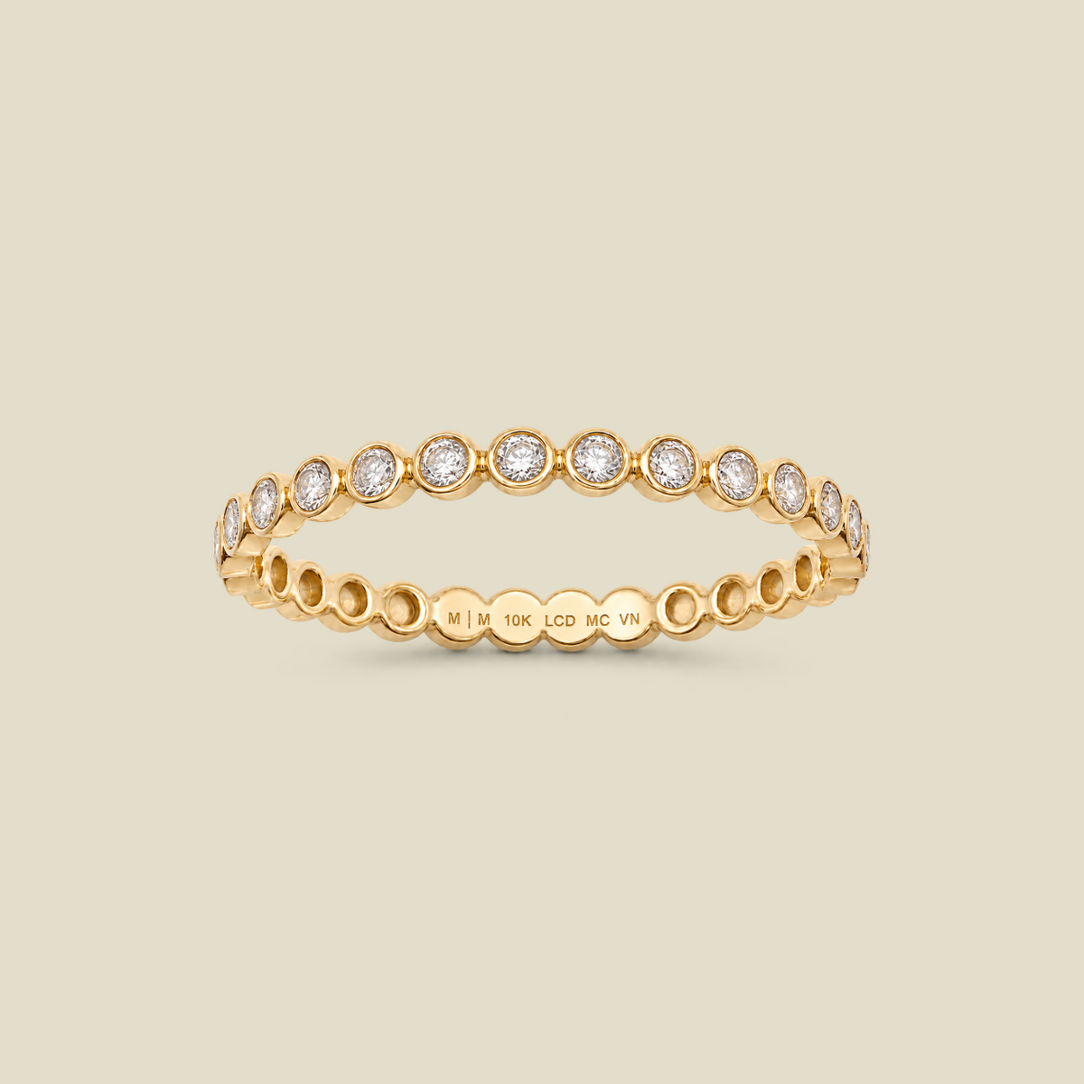 A delicate yellow gold eternity-style ring featuring a band of round-cut bezel-set diamonds, shown on a solid light beige background. 