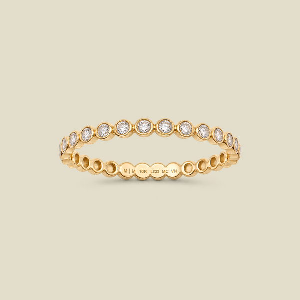 A delicate yellow gold eternity-style ring featuring a band of round-cut bezel-set diamonds, shown on a solid light beige background. 