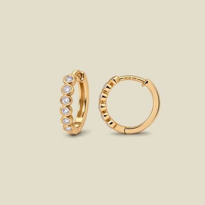 A pair of gold huggie hoop earrings, one shown from the front with six bezel-set round clear diamonds and the other from the side showing the hinge closure. The earrings are displayed on a solid light beige background.
