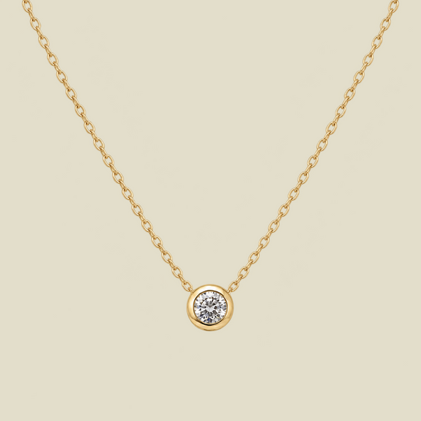 A delicate gold chain necklace with a small round bezel-set diamond pendant, displayed against a solid light beige background.