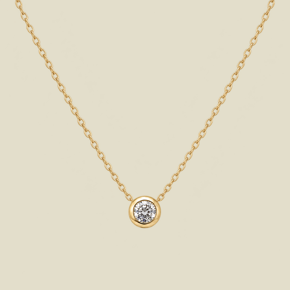 A delicate gold chain necklace with a small round bezel-set diamond pendant, displayed against a solid light beige background.