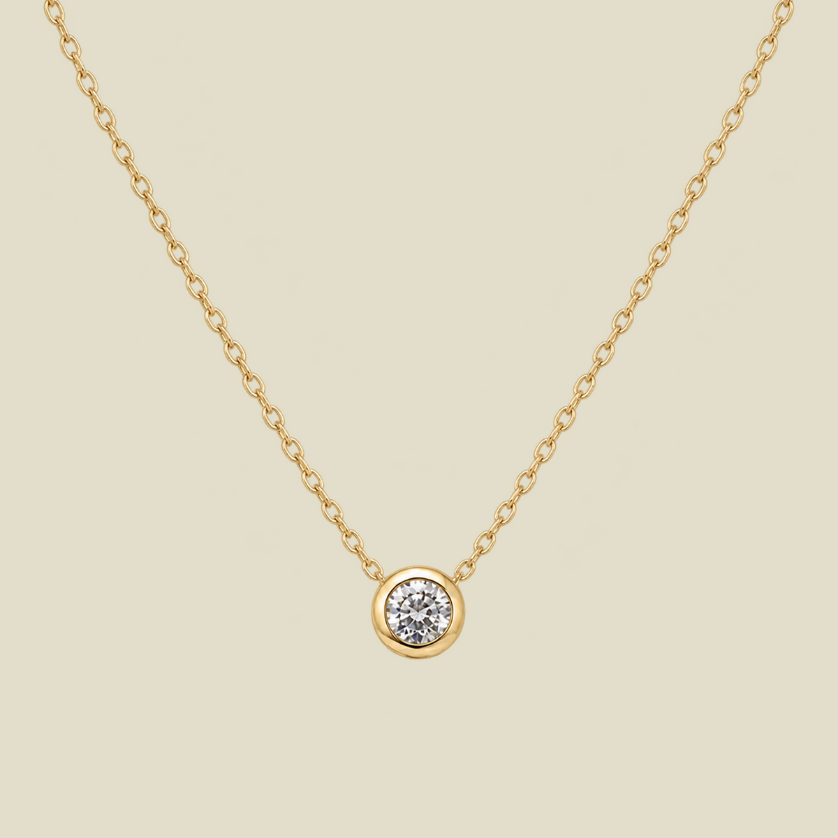 A delicate gold chain necklace with a small round bezel-set diamond pendant, displayed against a solid light beige background.