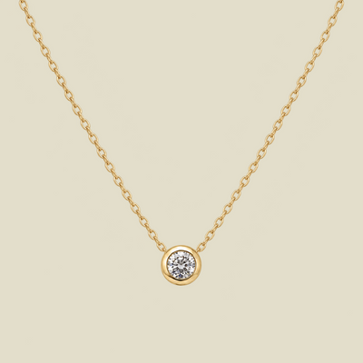 A delicate gold chain necklace with a small round bezel-set diamond pendant, displayed against a solid light beige background.