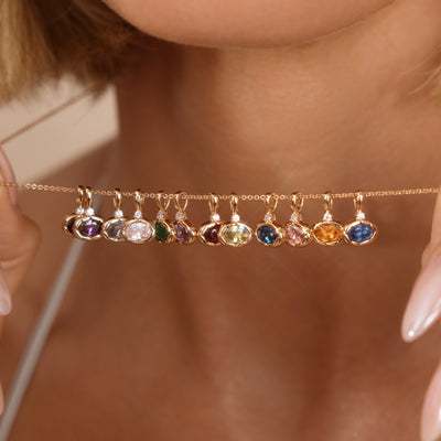 A close-up lifestyle shot of a model holding a delicate gold chain necklace with multiple small, colorful gemstone charms dangling horizontally across her neck.