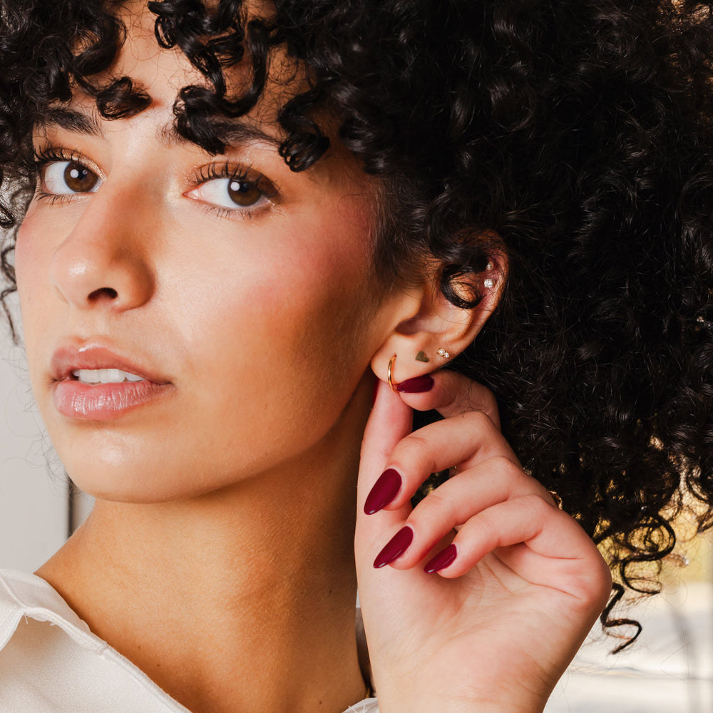 A close up lifestyle image of a model's face and ear, wearing a thin, small gold hoop and multiple gold studs in varying shapes and sizes.