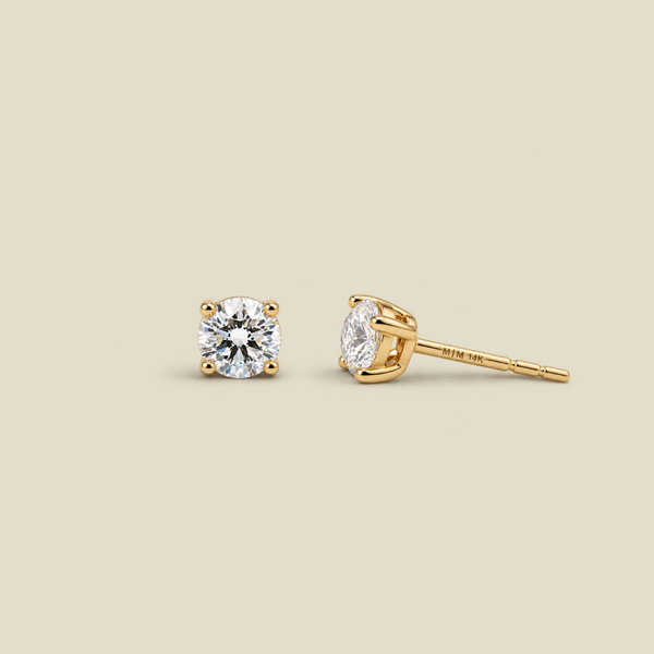 A pair of yellow four-prong gold stud earrings with round-cut diamond gemstones, shown on a solid light beige background. One earring is facing forward, and the other is turned to show the post and backing.