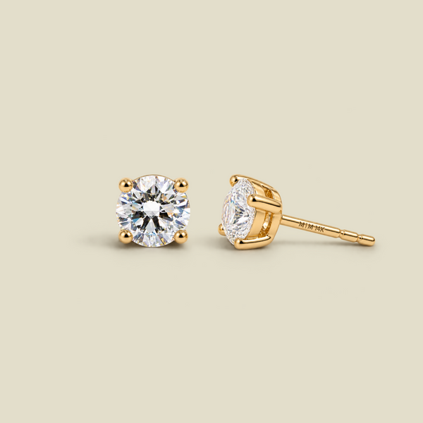 A pair of yellow four-prong gold stud earrings with round-cut diamond gemstones, shown on a solid light beige background. One earring is facing forward, and the other is turned to show the post and backing.