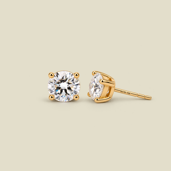 A pair of yellow four-prong gold stud earrings with round-cut diamond gemstones, shown on a solid light beige background. One earring is facing forward, and the other is turned to show the post and backing.