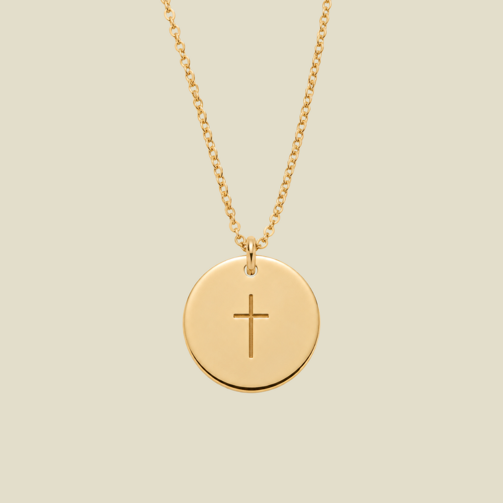 A gold round disc pendant necklace with a simple engraved cross in the center, suspended from a delicate gold chain against a solid light beige background.