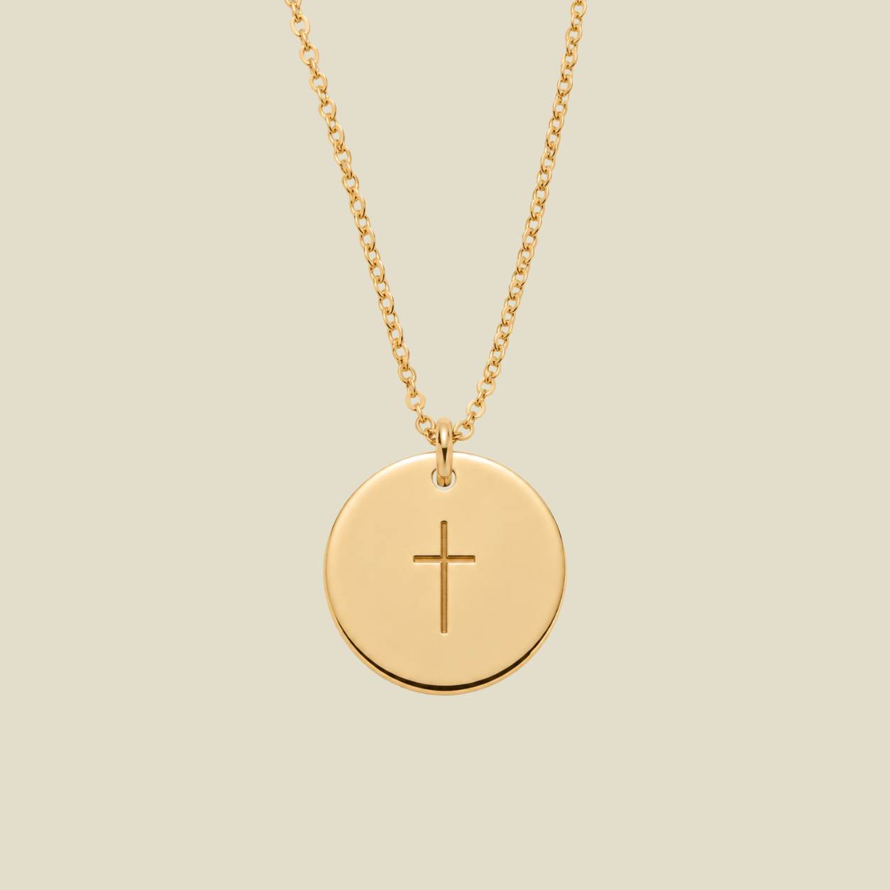 A gold round disc pendant necklace with a simple engraved cross in the center, suspended from a delicate gold chain against a solid light beige background.