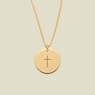 A gold round disc pendant necklace with a simple engraved cross in the center, suspended from a delicate gold chain against a solid light beige background.