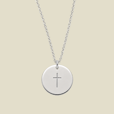 A silver round disc pendant necklace with a simple engraved cross in the center, suspended from a delicate silver chain against a solid light beige background.