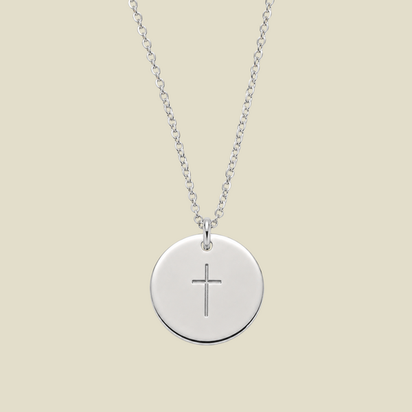 A silver round disc pendant necklace with a simple engraved cross in the center, suspended from a delicate silver chain against a solid light beige background.