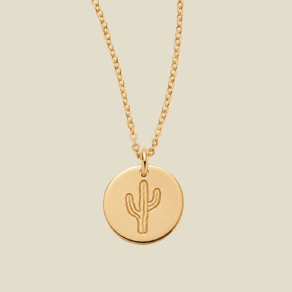 A gold chain necklace with a round gold disc pendant featuring an engraved cactus design, displayed against a solid light beige background.