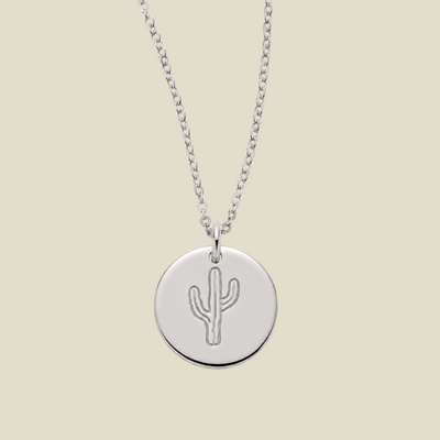 A silver round disc pendant necklace featuring an engraved cactus design, suspended from a delicate silver chain against a solid light beige background.