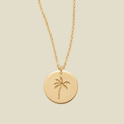 A gold disc pendant necklace featuring an engraved palm tree design, suspended from a delicate gold chain against a solid light beige background.