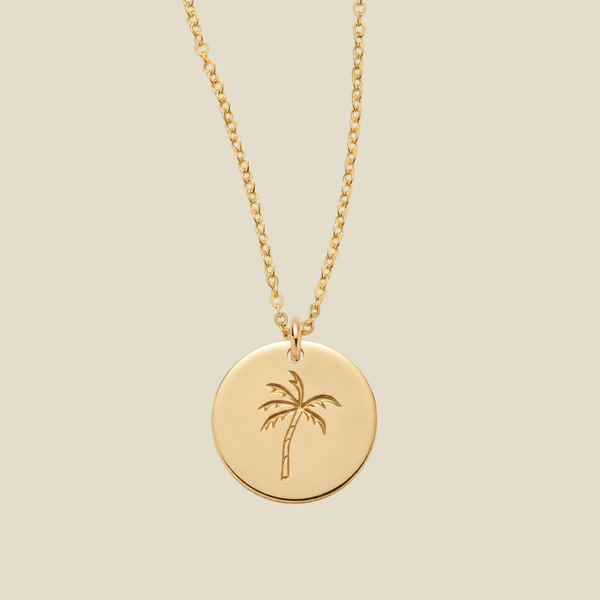 A gold disc pendant necklace featuring an engraved palm tree design, suspended from a delicate gold chain against a solid light beige background.