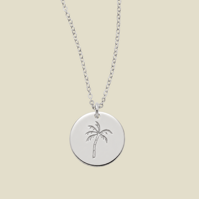 A silver round pendant necklace featuring an engraved palm tree design, suspended from a delicate silver chain against a solid light beige background.