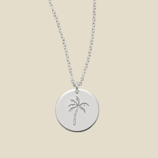 A silver round pendant necklace featuring an engraved palm tree design, suspended from a delicate silver chain against a solid light beige background.