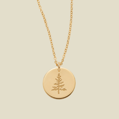 A gold chain necklace with a round gold disc pendant engraved with a pine tree, shown against a solid light beige background.