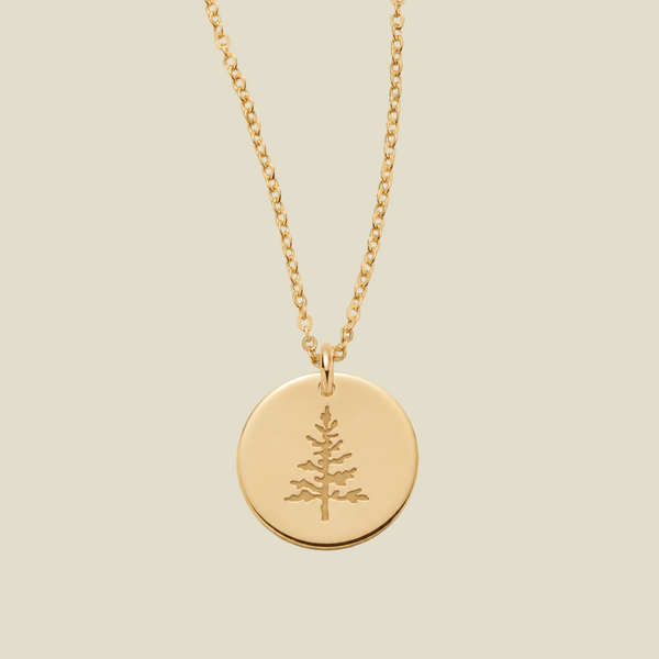 A gold chain necklace with a round gold disc pendant engraved with a pine tree, shown against a solid light beige background.