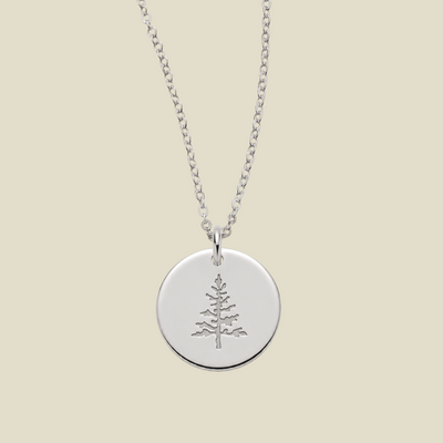 A silver round disc pendant necklace featuring an engraved pine tree design, suspended from a delicate silver chain against a solid light beige background.