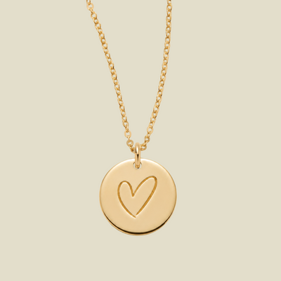 A gold chain necklace with a round disc pendant featuring a hand-drawn heart outline, shown against a solid light beige background.