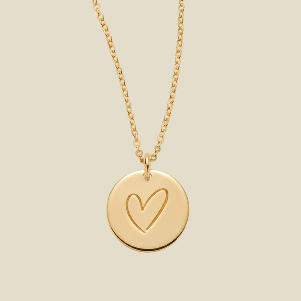 A gold chain necklace with a round disc pendant featuring a hand-drawn heart outline, shown against a solid light beige background.