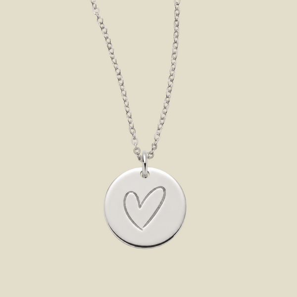 A silver round disc pendant necklace with a simple heart outline engraving, hanging from a delicate silver chain against a solid light beige background.
