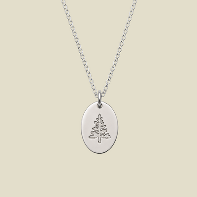 A silver oval pendant necklace featuring an engraved pine tree design, suspended from a delicate silver chain against a solid beige background.