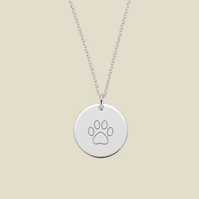 A silver round disc pendant necklace engraved with a paw print, hanging from a silver chain against a solid light beige background.