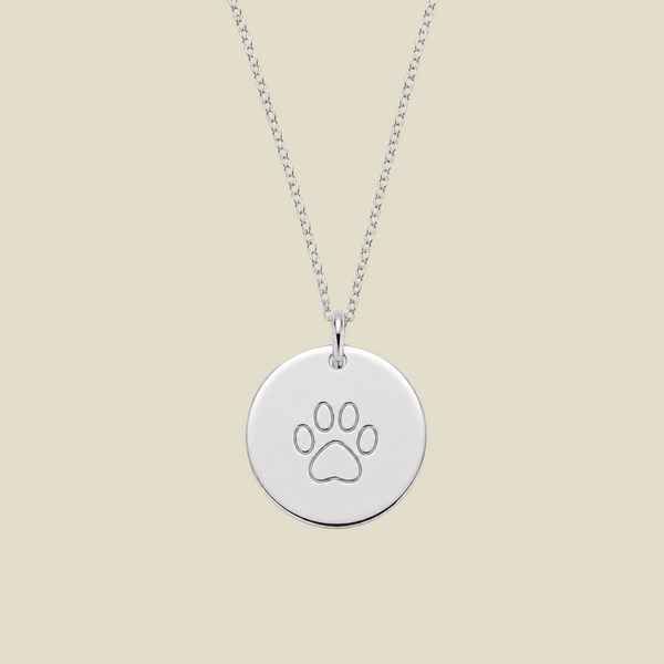A silver round disc pendant necklace engraved with a paw print, hanging from a silver chain against a solid light beige background.