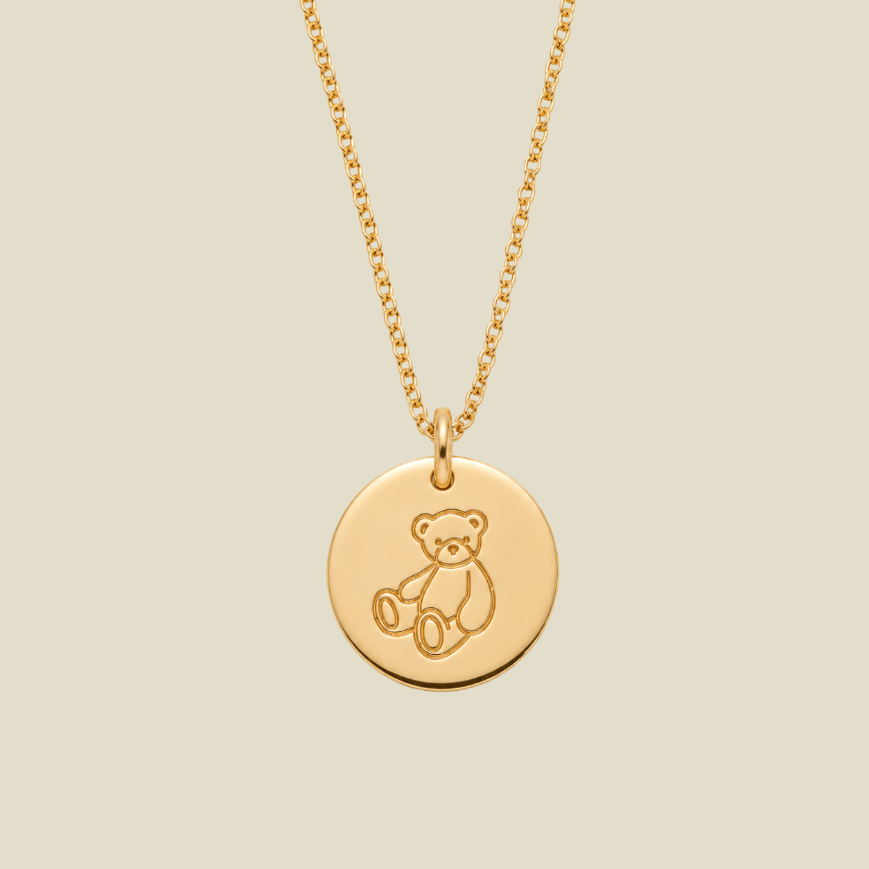 A gold disc pendant necklace featuring an engraved teddy bear design, suspended from a delicate gold chain against a solid light beige background.