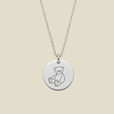 A silver round disc pendant necklace featuring a simple engraved teddy bear outline, suspended from a delicate silver chain against a solid light beige background.