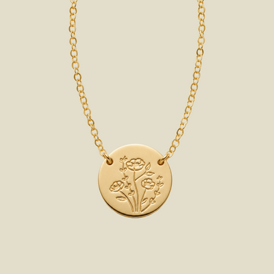 A gold chain necklace with a round gold disc pendant, attached to the chain at two points on the upper part of the disc, engraved with a floral design, shown against a solid light beige background.