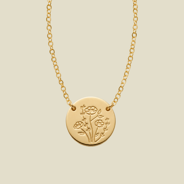A gold chain necklace with a round gold disc pendant, attached to the chain at two points on the upper part of the disc, engraved with a floral design, shown against a solid light beige background.