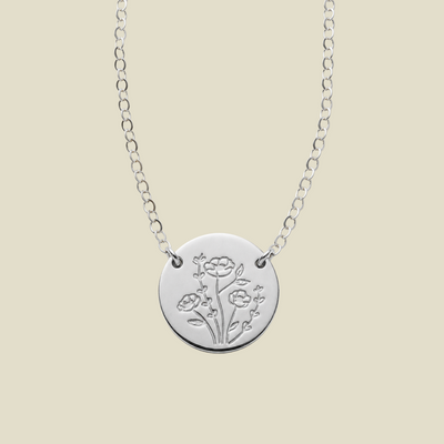 A silver chain necklace with a round disc pendant, attached at two points to the chain on the upper part of the disc, engraved with a floral design, shown against a solid light beige background.