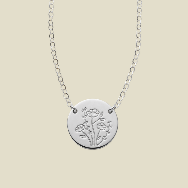 A silver chain necklace with a round disc pendant, attached at two points to the chain on the upper part of the disc, engraved with a floral design, shown against a solid light beige background.