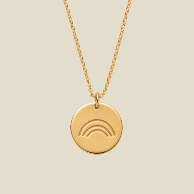 A gold round pendant necklace featuring an engraved minimalist rainbow design, suspended from a delicate gold chain against a solid light beige background.