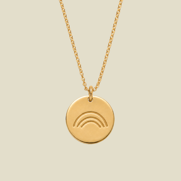 A gold round pendant necklace featuring an engraved minimalist rainbow design, suspended from a delicate gold chain against a solid light beige background.