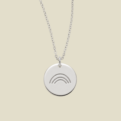 A silver round disc pendant necklace featuring an engraved rainbow design, suspended from a delicate silver chain against a solid beige background.