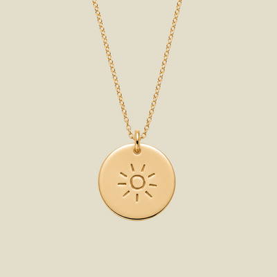 A gold circular pendant necklace featuring an engraved sun design, suspended from a delicate gold chain against a solid light beige background.