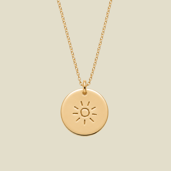 A gold circular pendant necklace featuring an engraved sun design, suspended from a delicate gold chain against a solid light beige background.