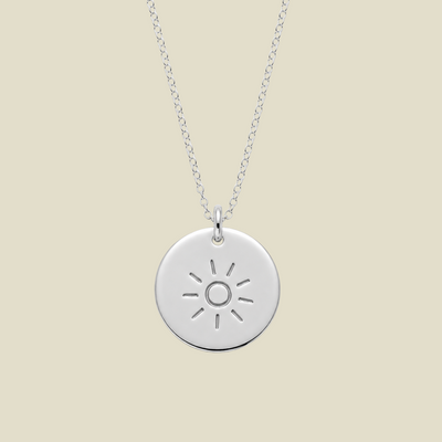 A silver round disc pendant necklace featuring a minimalistic sun design, suspended from a delicate silver chain against a solid light beige background.