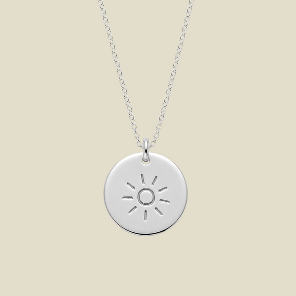 A silver round disc pendant necklace featuring a minimalistic sun design, suspended from a delicate silver chain against a solid light beige background.