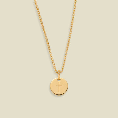 A gold chain necklace with a round gold disc pendant engraved with a cross, displayed against a solid light beige background.