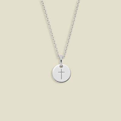 A silver round disc pendant necklace with a simple cross engraving, suspended from a delicate silver chain against a solid beige background.