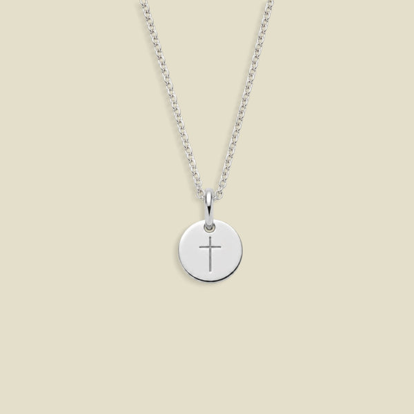 A silver round disc pendant necklace with a simple cross engraving, suspended from a delicate silver chain against a solid beige background.