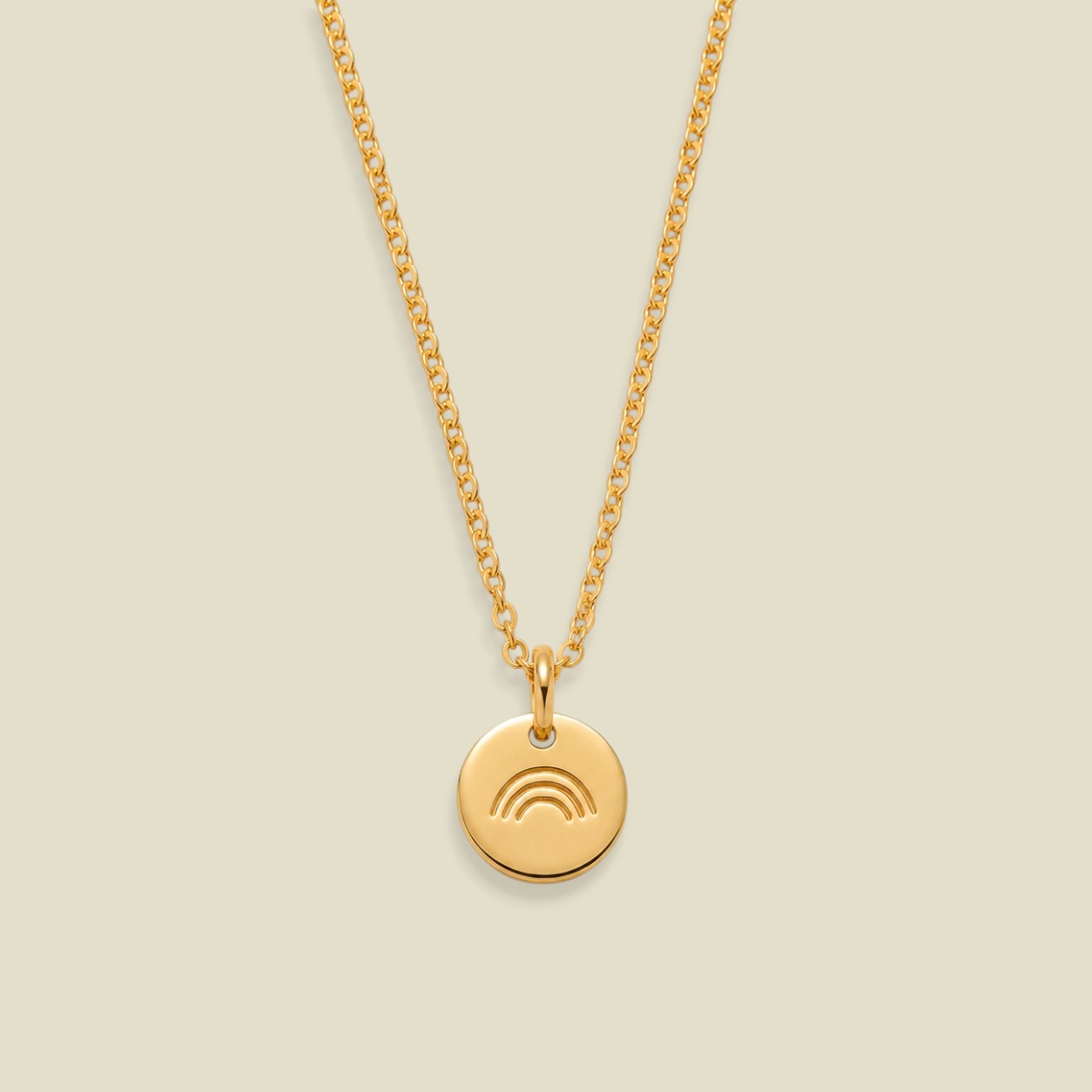 A gold chain necklace with a small round gold pendant featuring an engraved rainbow design, displayed against a solid light beige background.