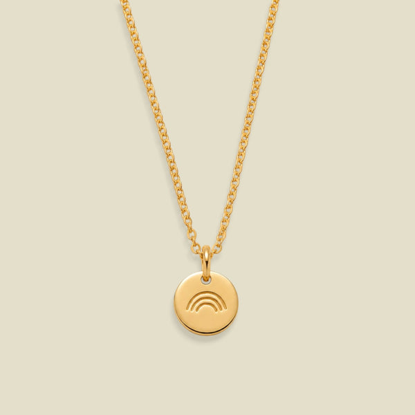 A gold chain necklace with a small round gold pendant featuring an engraved rainbow design, displayed against a solid light beige background.
